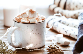 Un chocolat chaud avec des guimauves dans une tasse en céramique blanche entourée de choses d'hiver sur une table en bois.