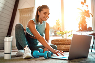 Une femme sportive en vêtements de sport est assise sur le sol avec des haltères et une bouteille d'eau et utilise un ordinateur portable