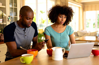 Un couple noir regarde leur ordinateur et leur téléphone tout en prenant leur petit-déjeuner. 