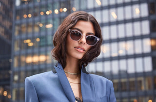 Une jeune femme portant des lunettes de soleil Maui Jim transparentes et une veste bleue devant un gratte-ciel.