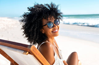 Portrait d'une jeune femme noire portant des lunettes de soleil qui se repose sur un transat en bois à la plage tout en regardant la caméra avec un sourire