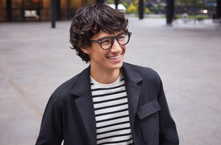 Jeune homme souriant portant des lunettes Tom Ford et une veste noire par-dessus un t-shirt rayé noir et blanc.