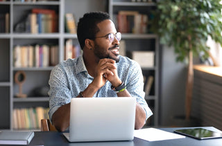 Un homme noir avec des lunettes rondes est assis à son bureau devant son ordinateur portable. Il regarde à l'extérieur par une fenêtre.