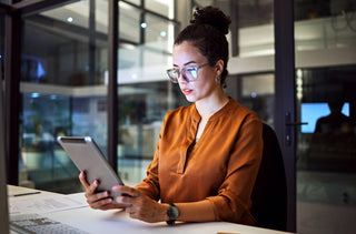 Une femme avec des lunettes et un chemisier orange consulte sa tablette dans son bureau.