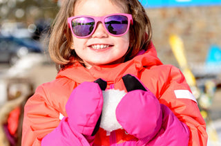 Une jeune fille portant un habit de neige rose et orange avec des lunettes de soleil roses tient une boule de neige dans ses mains. 